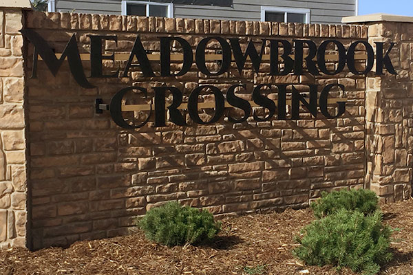 Stone sign with black letters reading "MEADOWBROOK CROSSING," surrounded by small bushes.