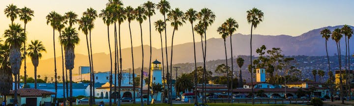 A scenic view featuring palm trees, mountains, and buildings at sunset, capturing a tranquil coastal atmosphere.
