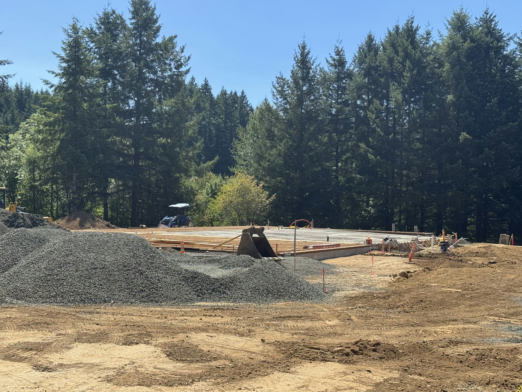 Construction site with gravel, dirt, a tractor, and a forest background on a sunny day.