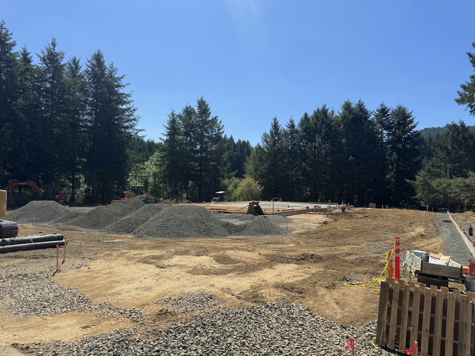 Construction site with gravel piles, machinery, and forested background.