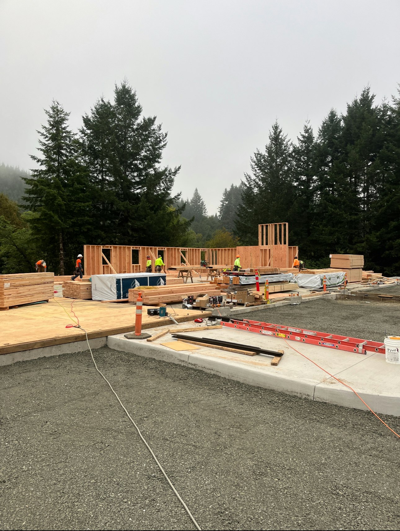 Construction site with workers building a wooden structure, surrounded by trees.