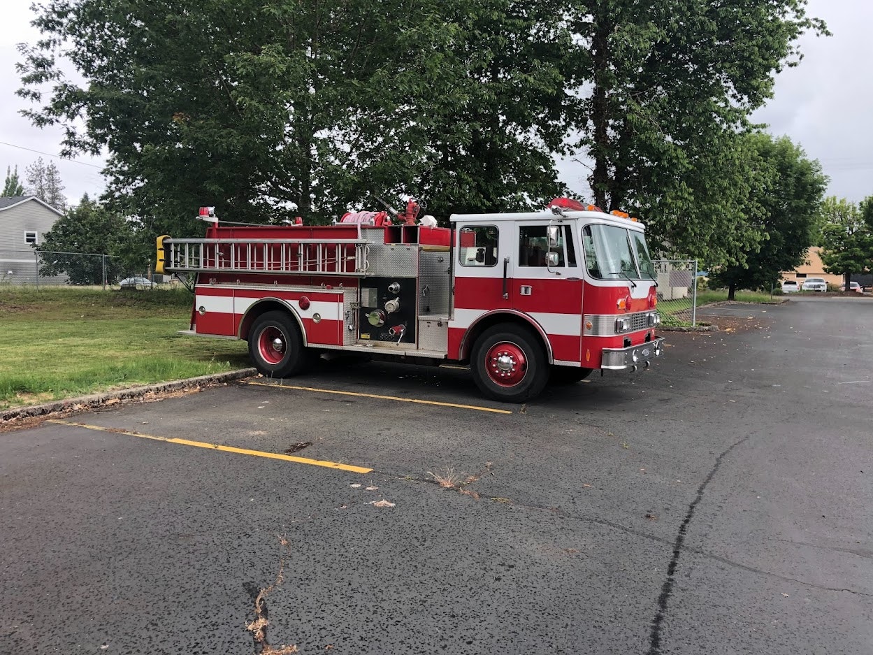 A red fire truck parked on a wet asphalt surface, with trees and houses in the background.