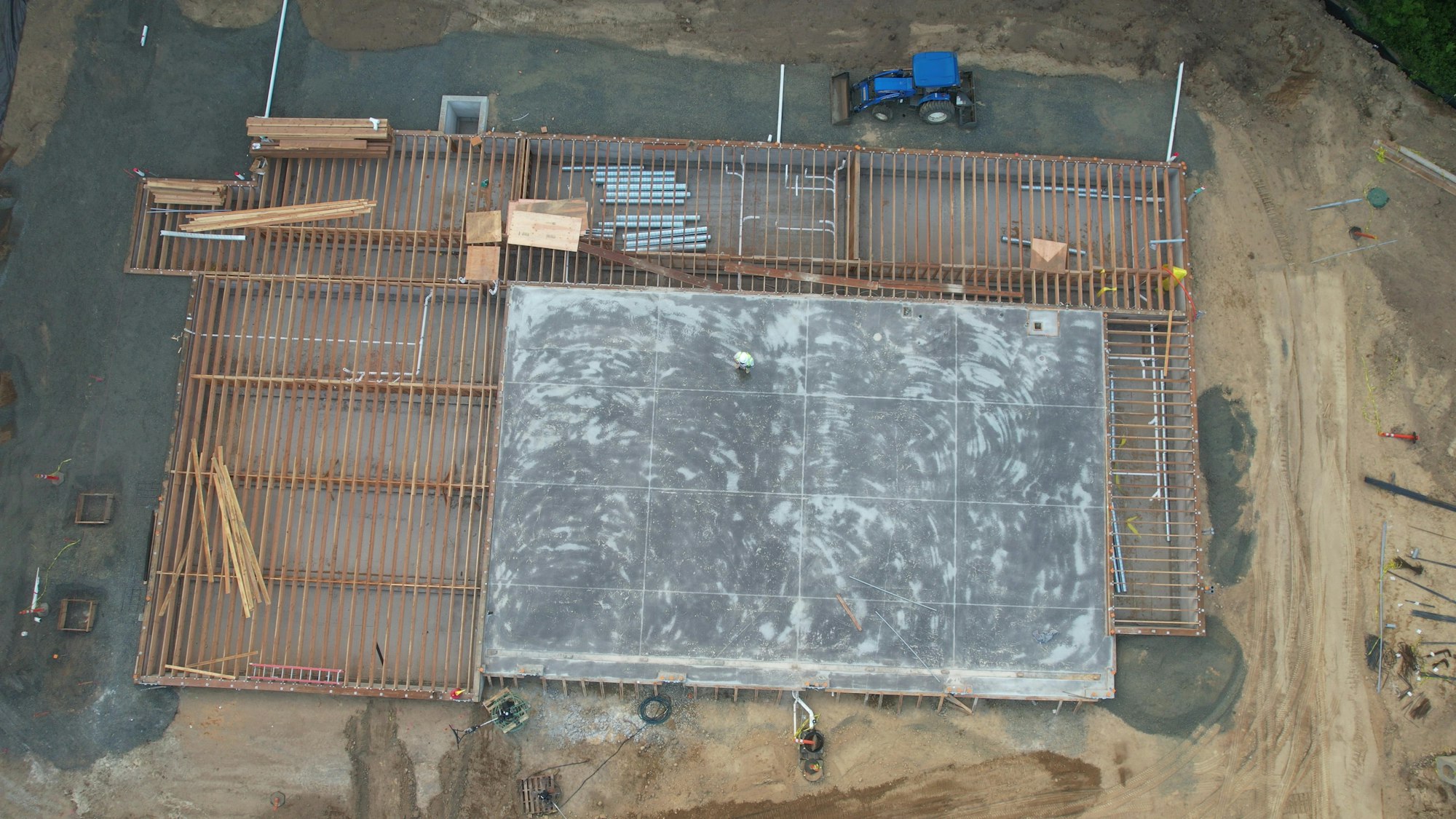 Aerial view of a construction site with wood framing, concrete slab, and a blue tractor nearby.