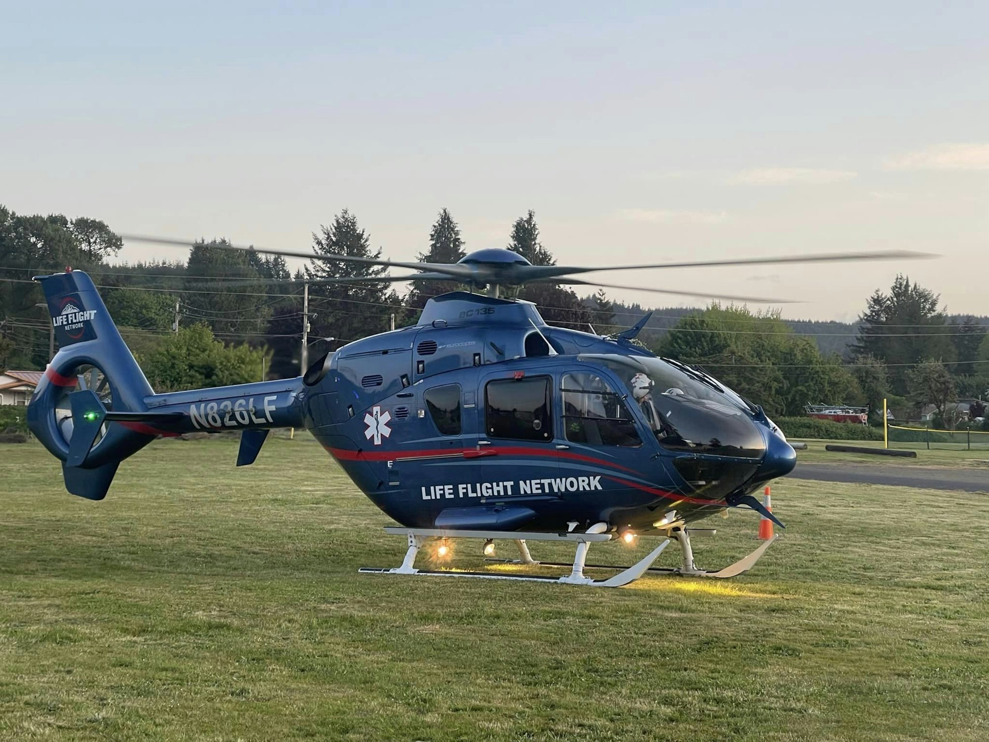 A medical helicopter on a grassy field at dusk, rotors spinning, no people in view.