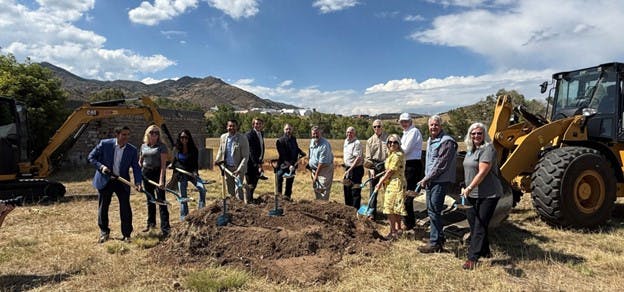 A group of people at a groundbreaking ceremony, holding shovels, with construction equipment in the background.