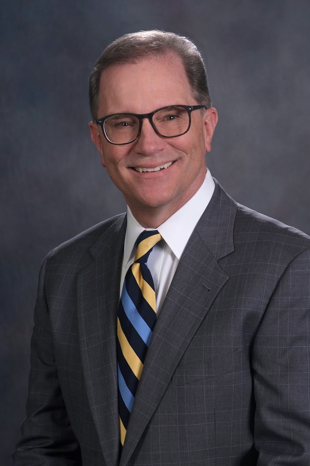 A smiling man wearing glasses, a suit, a striped tie, and a white shirt against a grey backdrop.