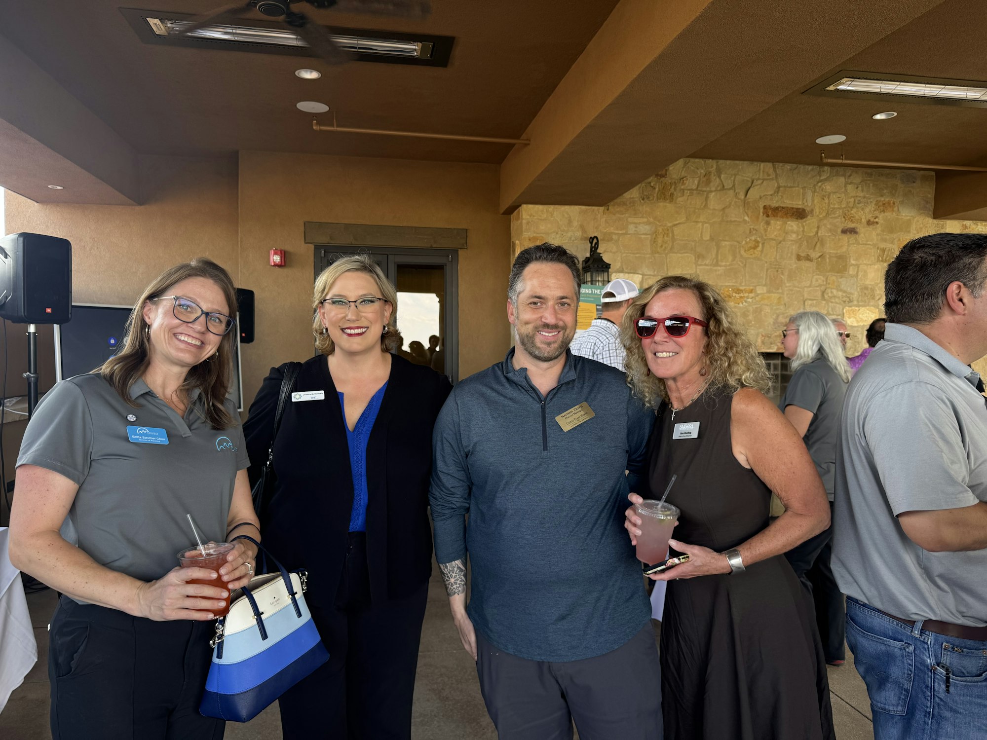 Four people smiling at an event, two holding drinks. Indoors with a stone wall background.