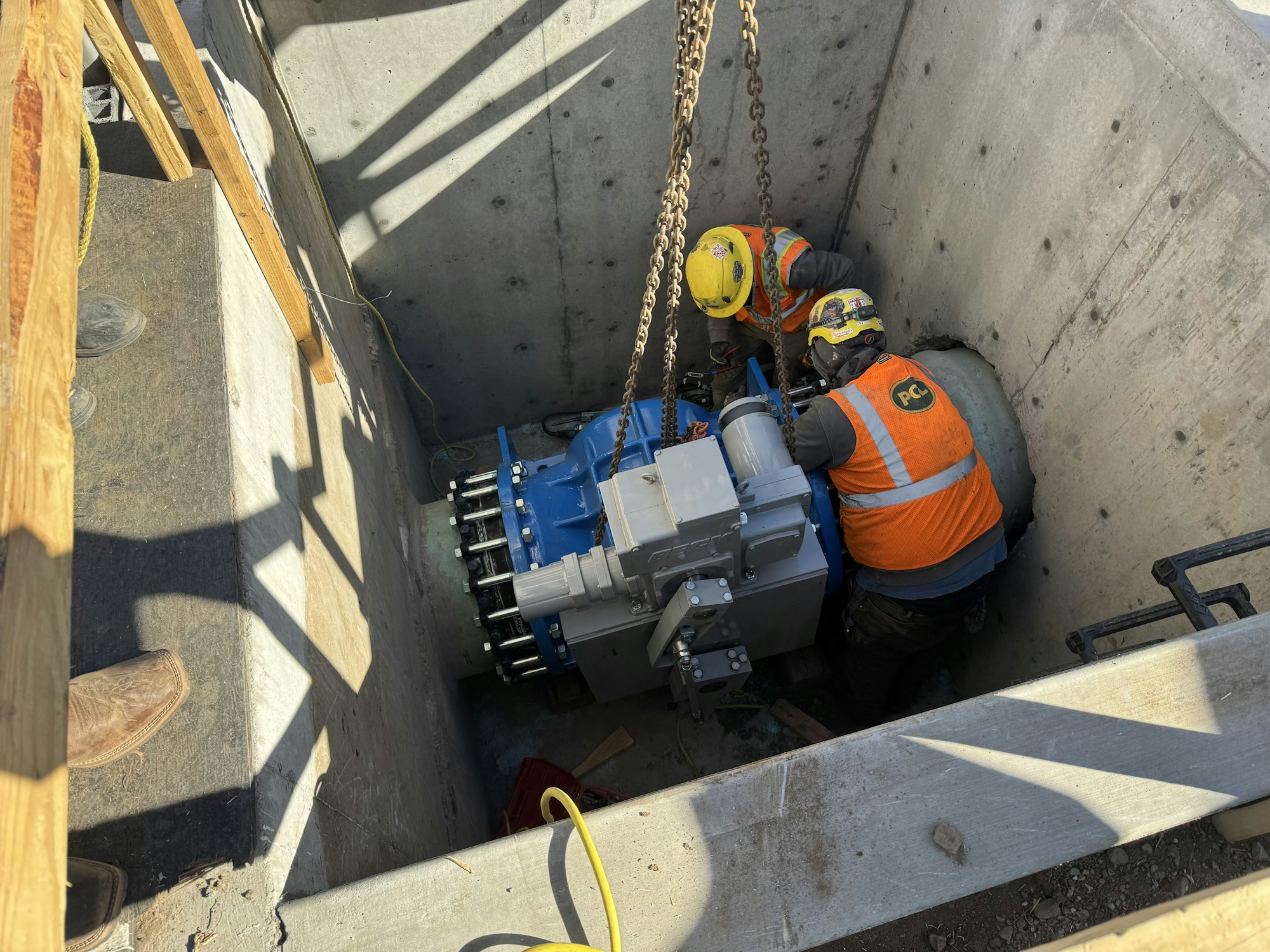 Two workers wearing safety gear are installing machinery at the bottom of a concrete pit.