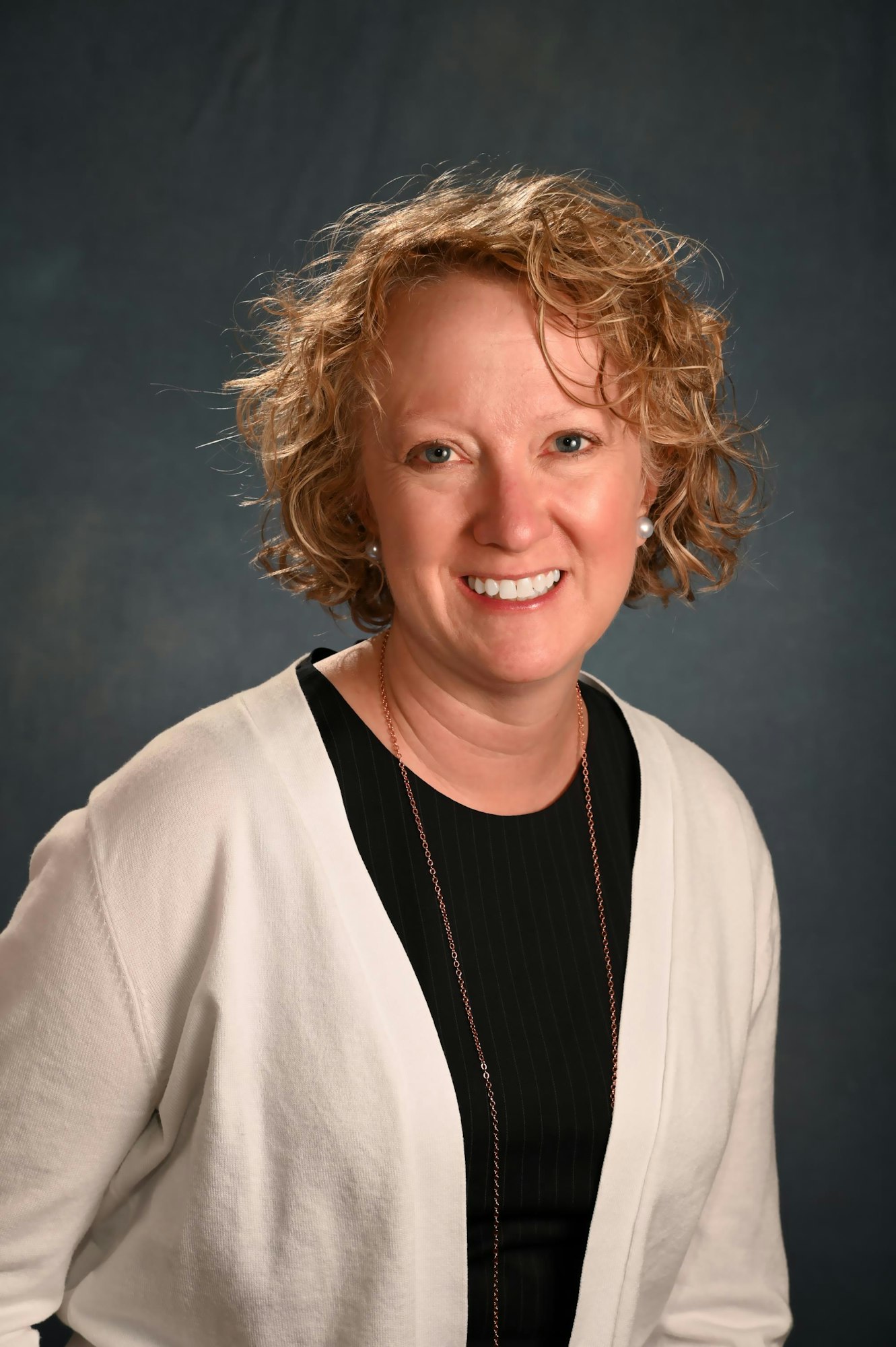 A smiling woman with curly hair wearing a white cardigan, black top, and necklace against a mottled backdrop.