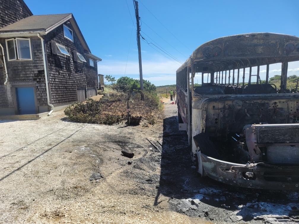 A burned-out bus next to a house on a sunny day.