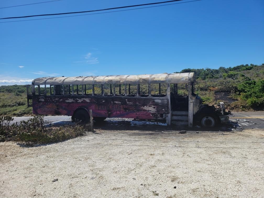 A burnt-out bus on a rural road, surrounded by vegetation under a clear blue sky.