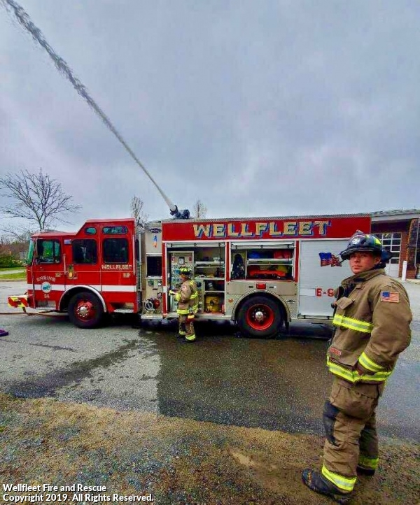 Firefighters operating a hose from a fire truck labeled "WELLFLEET."