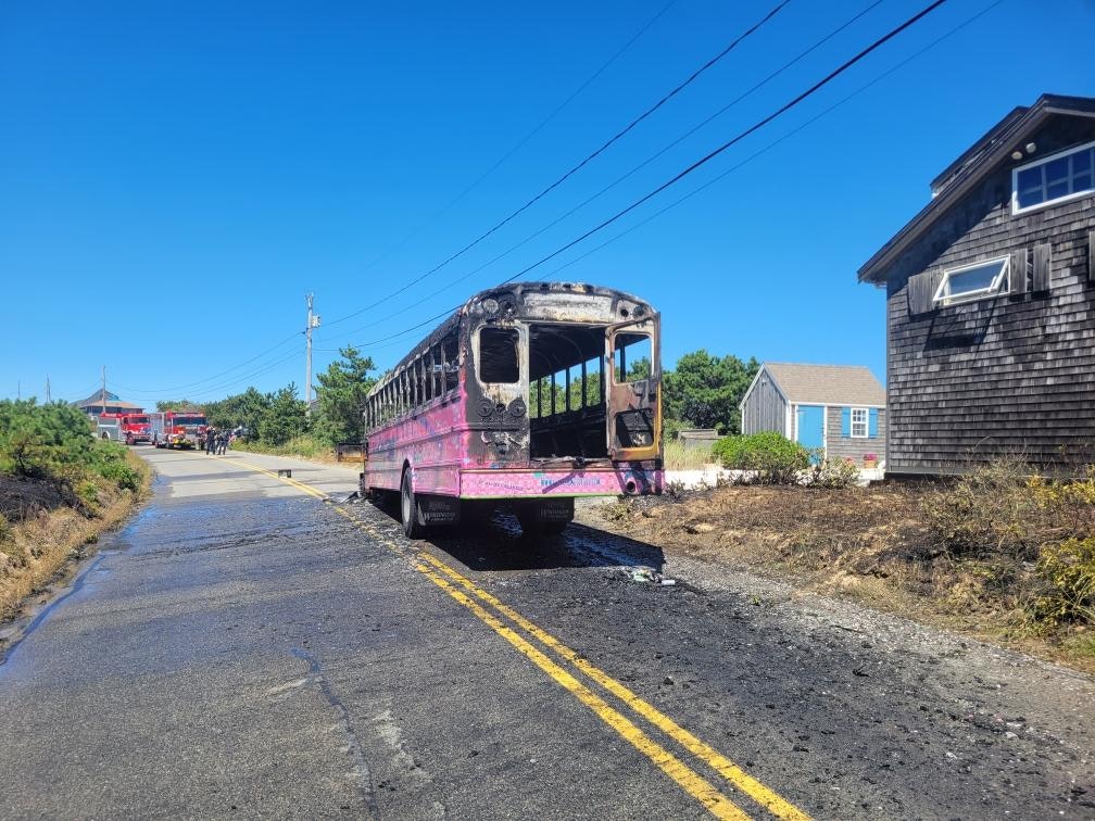 A burnt-out bus is on a road. Fire trucks and firefighters are in the distance. Nearby houses are visible under a clear blue sky.