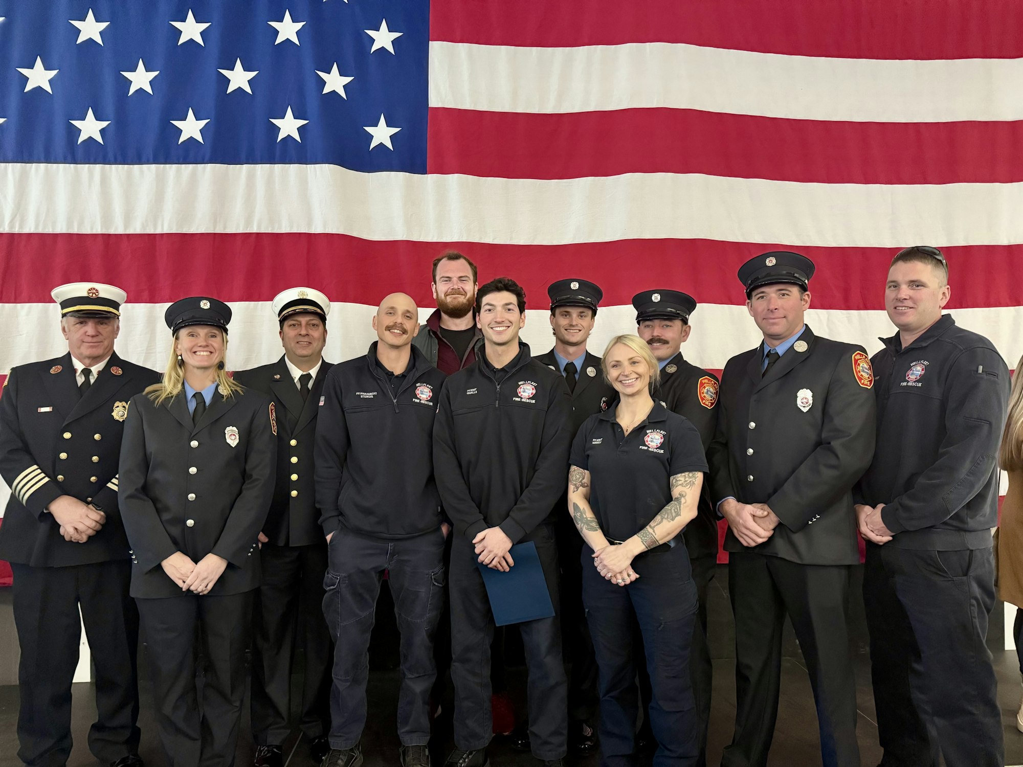 A group of fire service personnel poses in front of a large American flag, showcasing their pride and teamwork.
