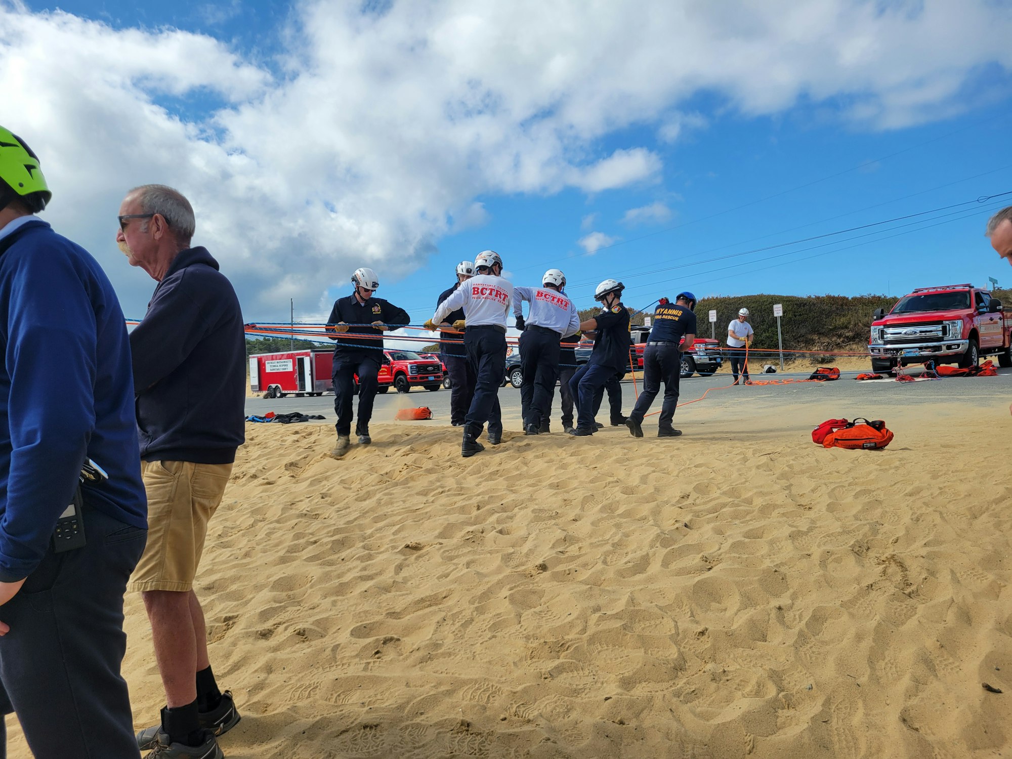 Emergency responders on a sandy area with equipment, fire trucks, and rescue operation setup.