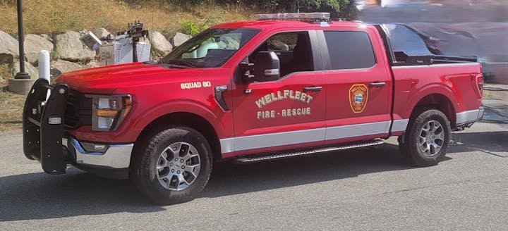 A red fire-rescue truck labeled "Wellfleet" with emergency lights and a push bar, parked on a sunny day.