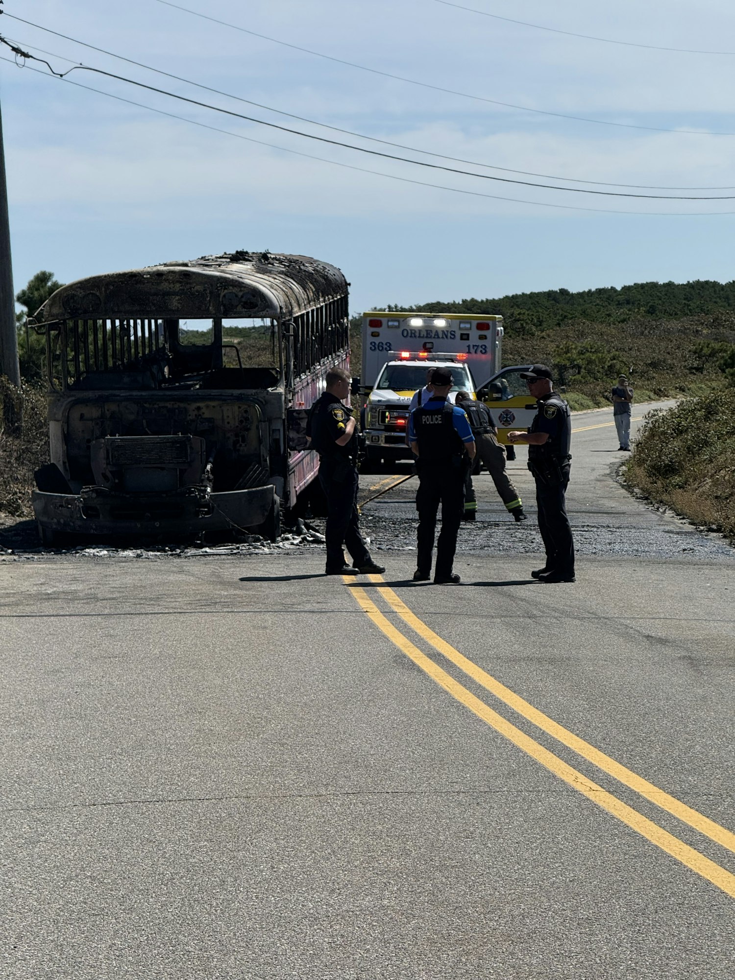 Burnt vehicle with emergency personnel near an ambulance on a road.