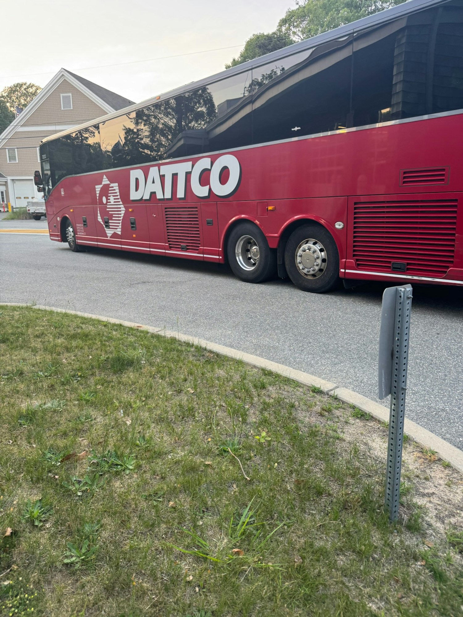 A red DATTCO bus parked on a street near a house, with grass and a signpost in the foreground.