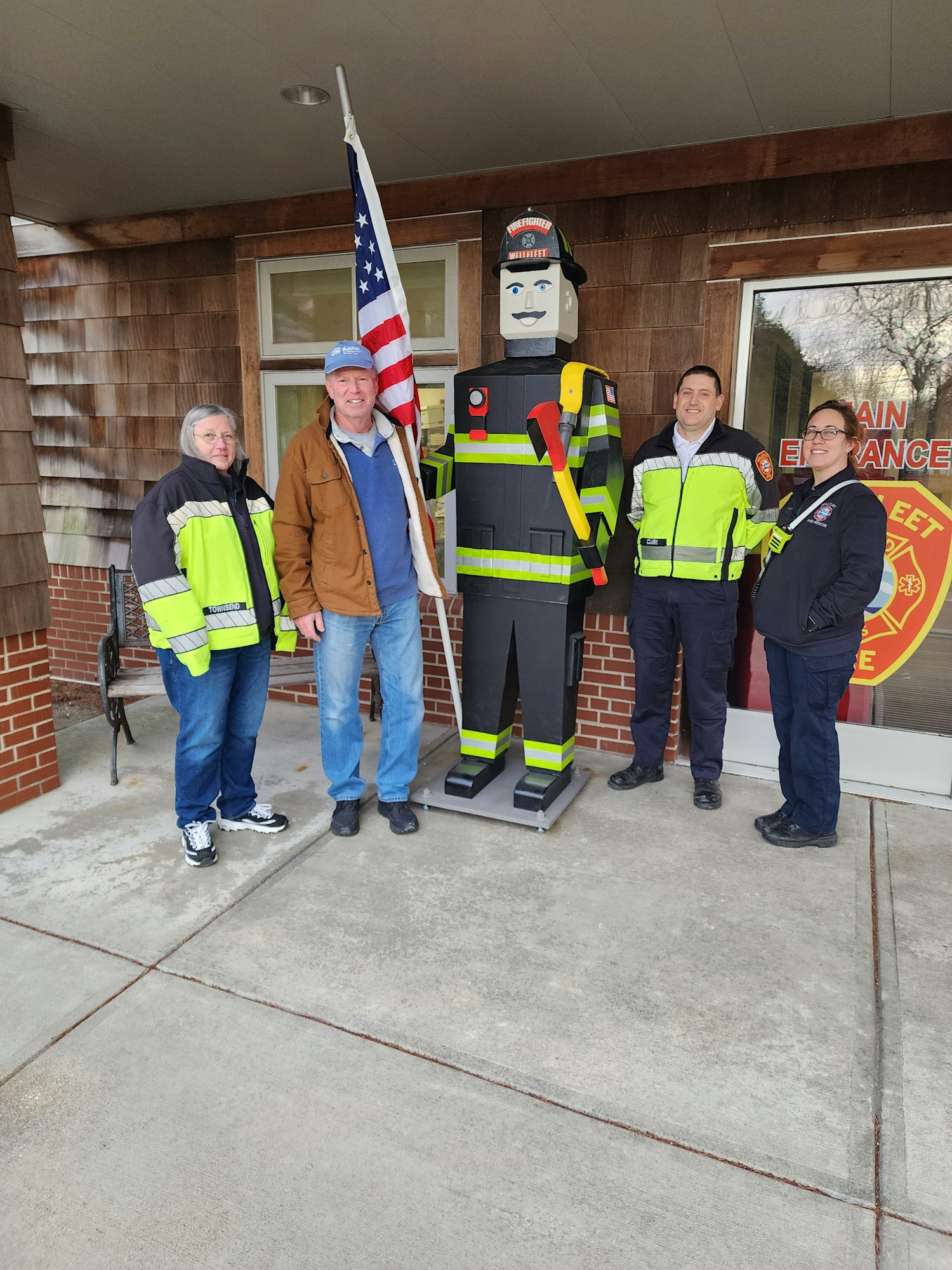 Four people standing outside a building; one holds a U.S. flag, next to a large firefighter cutout.