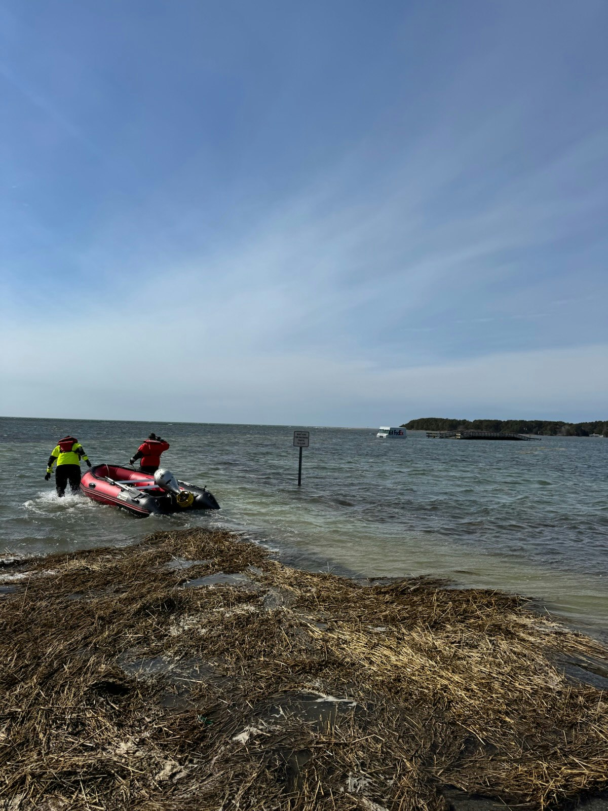 Two people in bright jackets push a red inflatable boat into the water near a sign, with a distant boat and dock in the background.