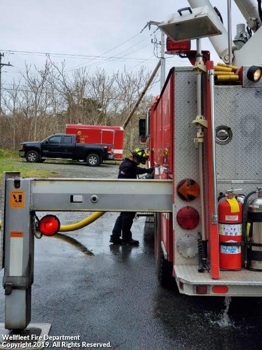 A firefighter works near a fire truck with hoses and equipment, with another vehicle in the background.
