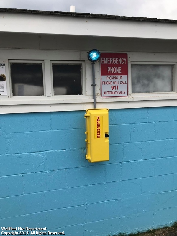 A yellow emergency phone box on a blue wall with a sign indicating that lifting the phone dials 911 automatically, and a blue light above.