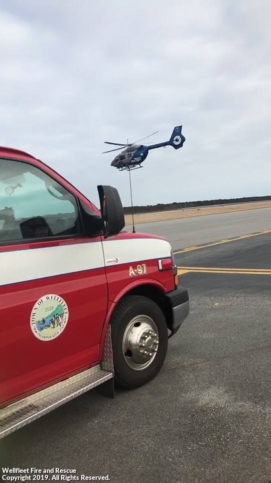 A helicopter in flight behind a partial view of a fire department vehicle on an airstrip.