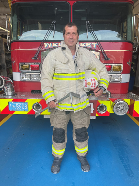 A firefighter in full gear stands in front of a fire engine.