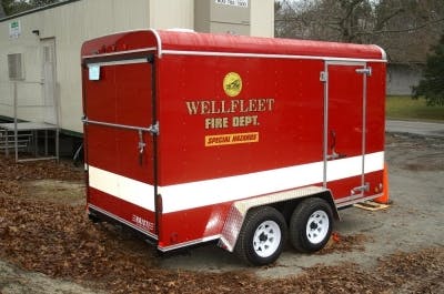 A red trailer with "WELLFLEET FIRE DEPT. SPECIAL HAZARDS" written on it, parked on dirt ground.