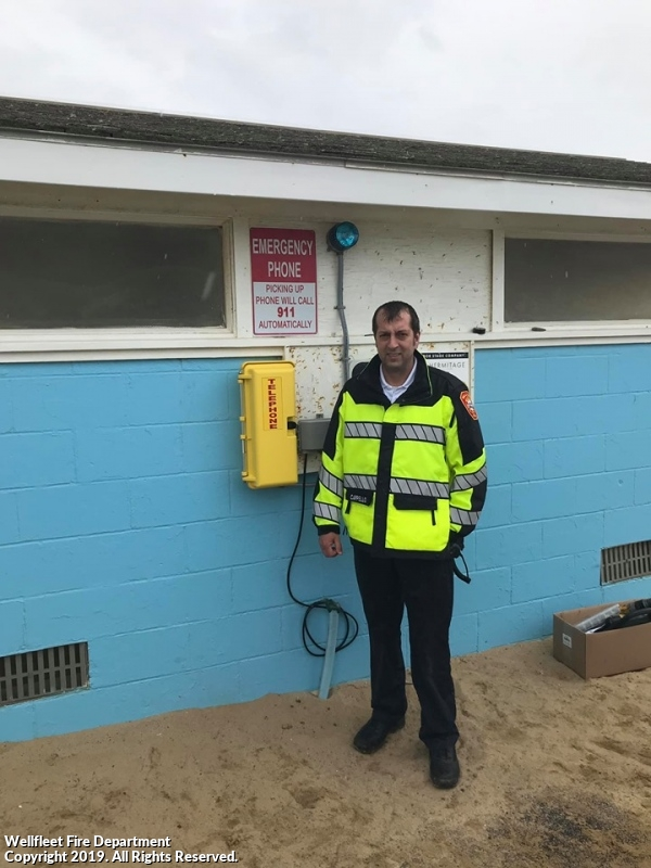 Man in high-visibility jacket standing next to emergency phone on building wall.