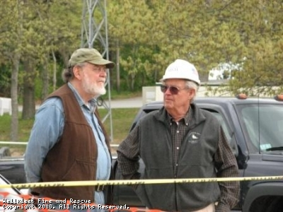 Two men conversing, one in a cap and the other in a hard hat, with vehicles in the background.
