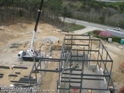 A construction site with a crane lifting materials and a partially built steel structure, trees, and a road in the background.