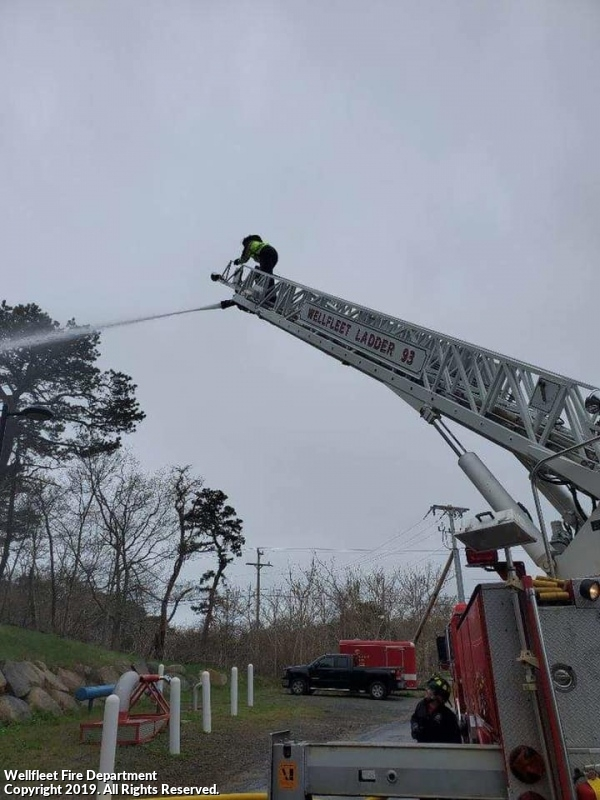 Firefighter on extended ladder spraying water, fire truck below, outdoor setting.