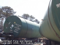 Green cylindrical water tank with "Wellfleet Fire and Rescue" text, mounted on supports, trees in background.