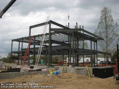 Steel framework of a building under construction with a crane, cloudy sky, and construction materials around.