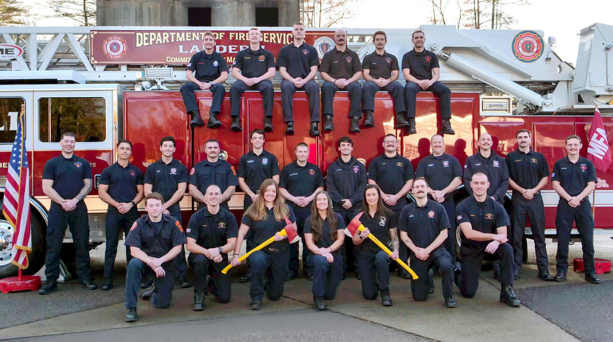 A group of firefighters poses in front of a fire truck, showcasing teamwork and camaraderie. Tools included: axes.