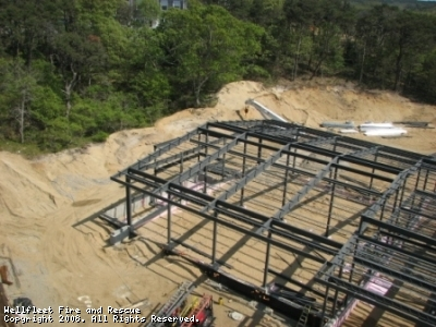 Construction site with metal framework of a building in progress, surrounded by dirt and vegetation, under daylight.