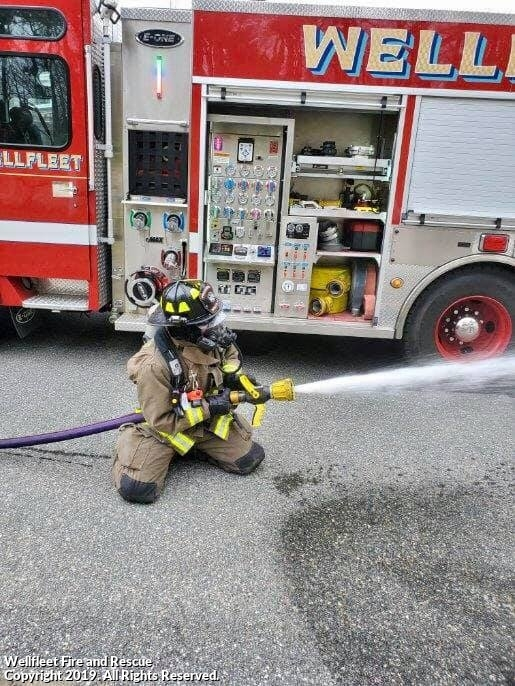 A firefighter kneels, operating a hose with a powerful stream of water, next to a fire truck labeled "WELLFLEET".