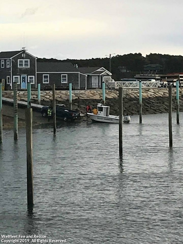 A pickup truck submerged near a dock, with people in a boat beside it, wooden poles, and buildings in the background.