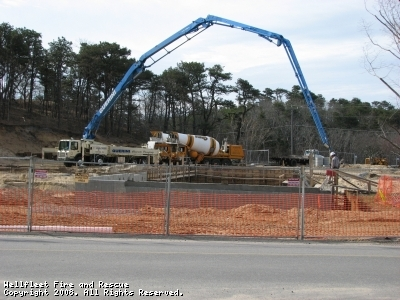 Construction site with a concrete pump truck, cement mixer, and barriers, under a cloudy sky.