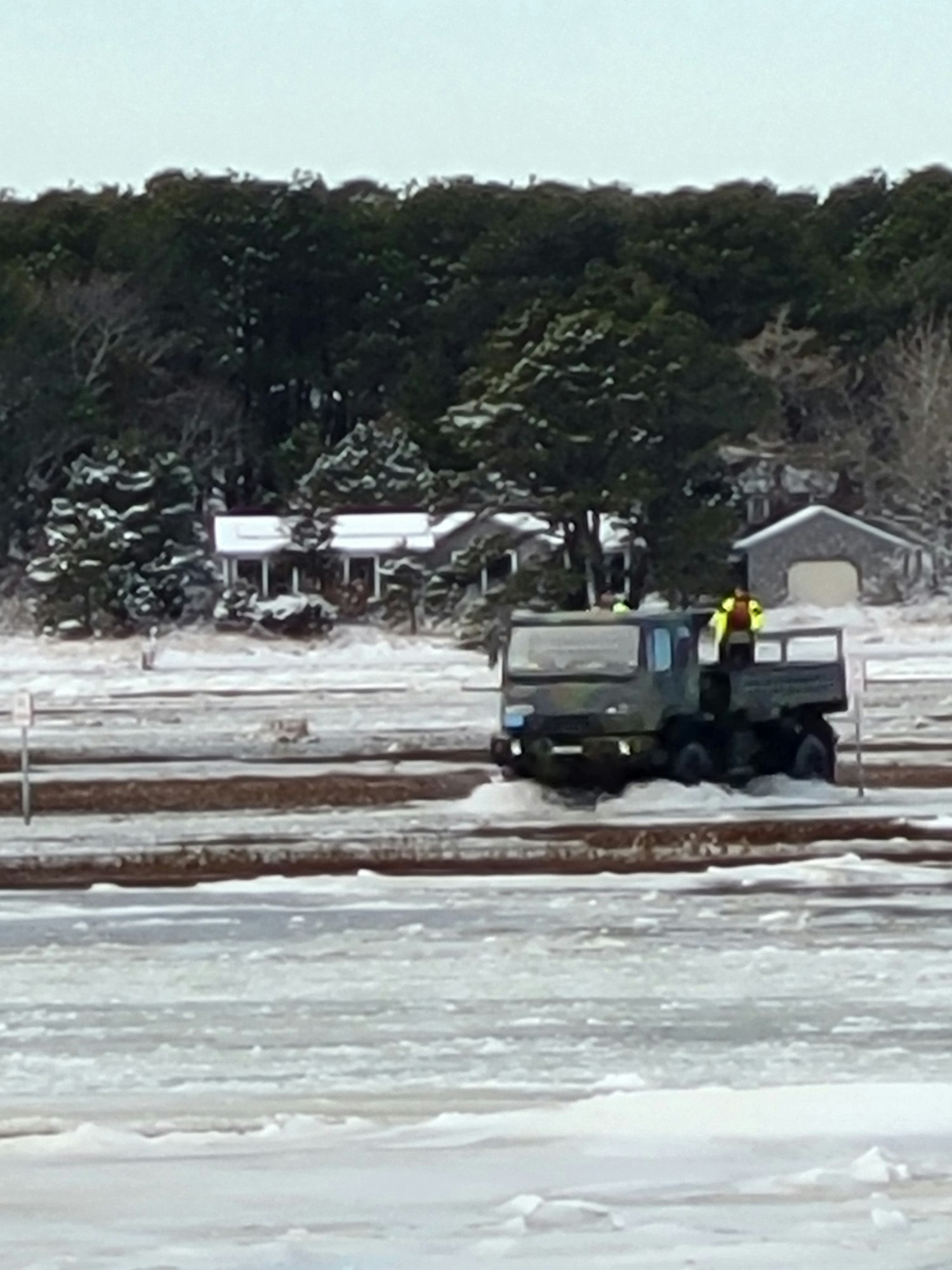 A vehicle is working on icy terrain near trees and buildings, likely clearing snow or performing maintenance.