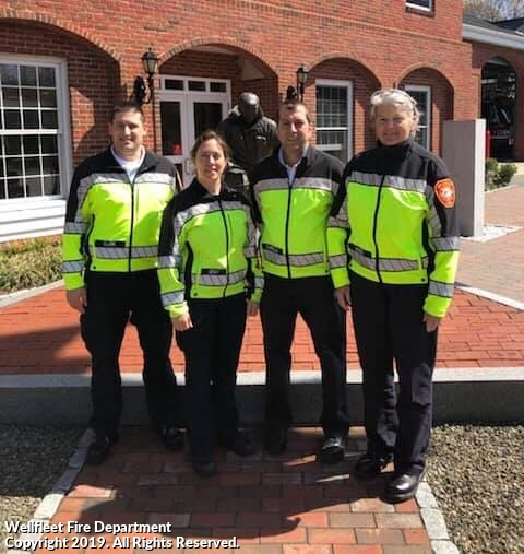 Four firefighters in high-visibility jackets stand in front of a brick building.