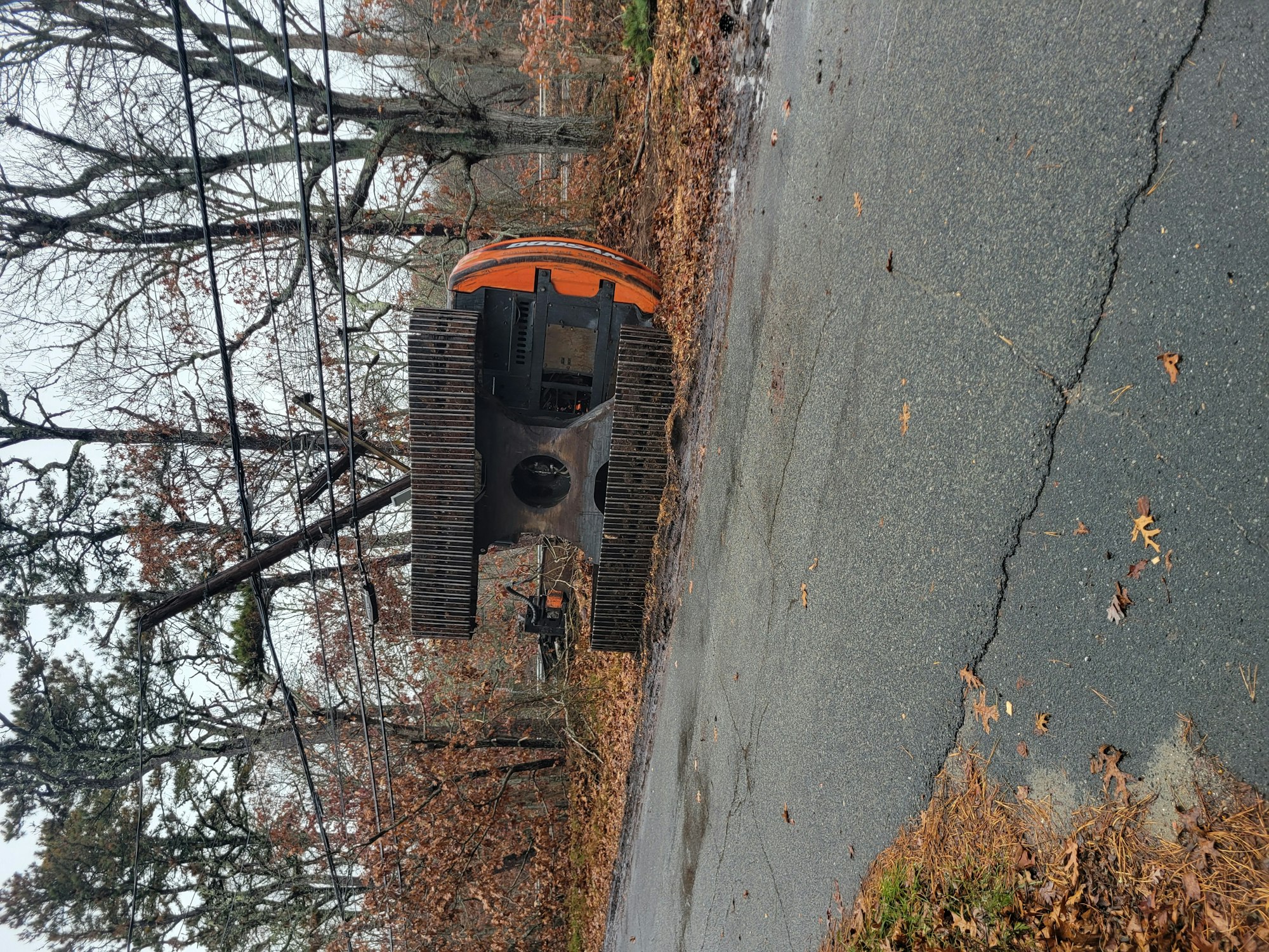 The image shows a large piece of construction equipment on a road surrounded by trees and fallen leaves, with a cloudy sky.