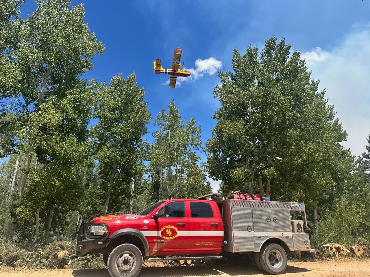 Red fire truck on a dirt road with trees in the background; yellow firefighting plane flying overhead against a blue sky.