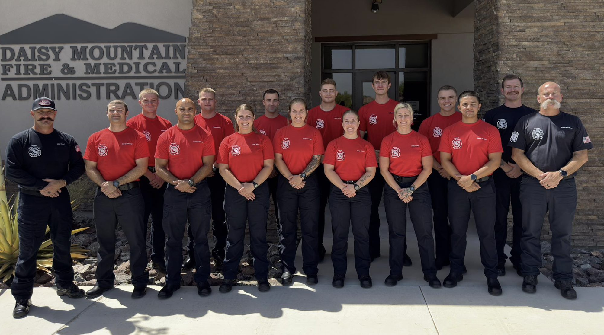 A group of people, likely firefighters, in red and black uniforms, standing in front of a building labeled "Daisy Mountain Fire & Medical Administration."