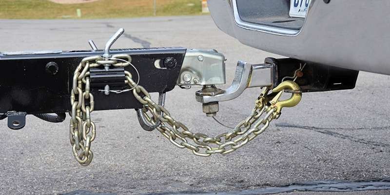 A trailer hitch with safety chains connects a vehicle to a trailer on a paved surface.