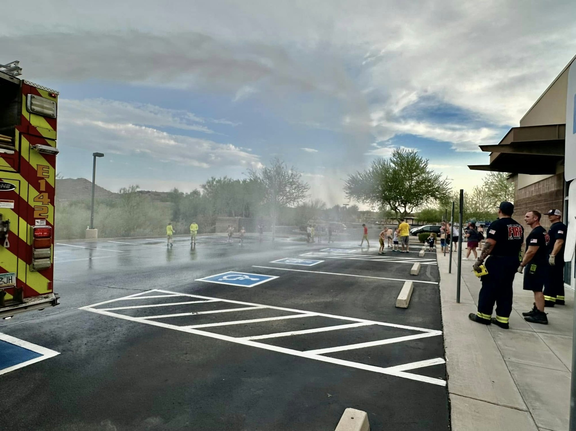 Firefighters watch as children play in water from a fire hose in a parking lot.