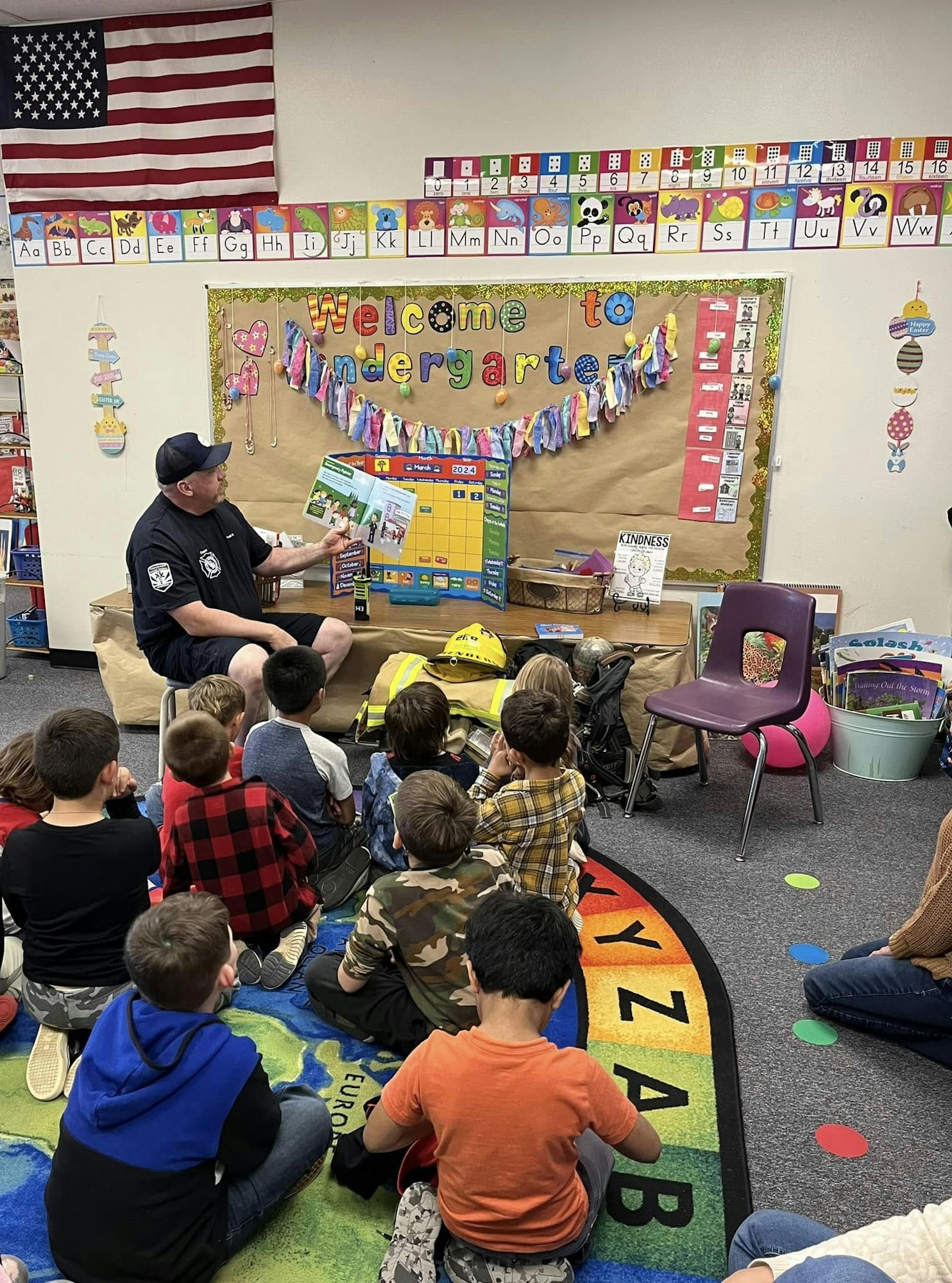A man reads to children in a kindergarten classroom with colorful decor and an American flag.