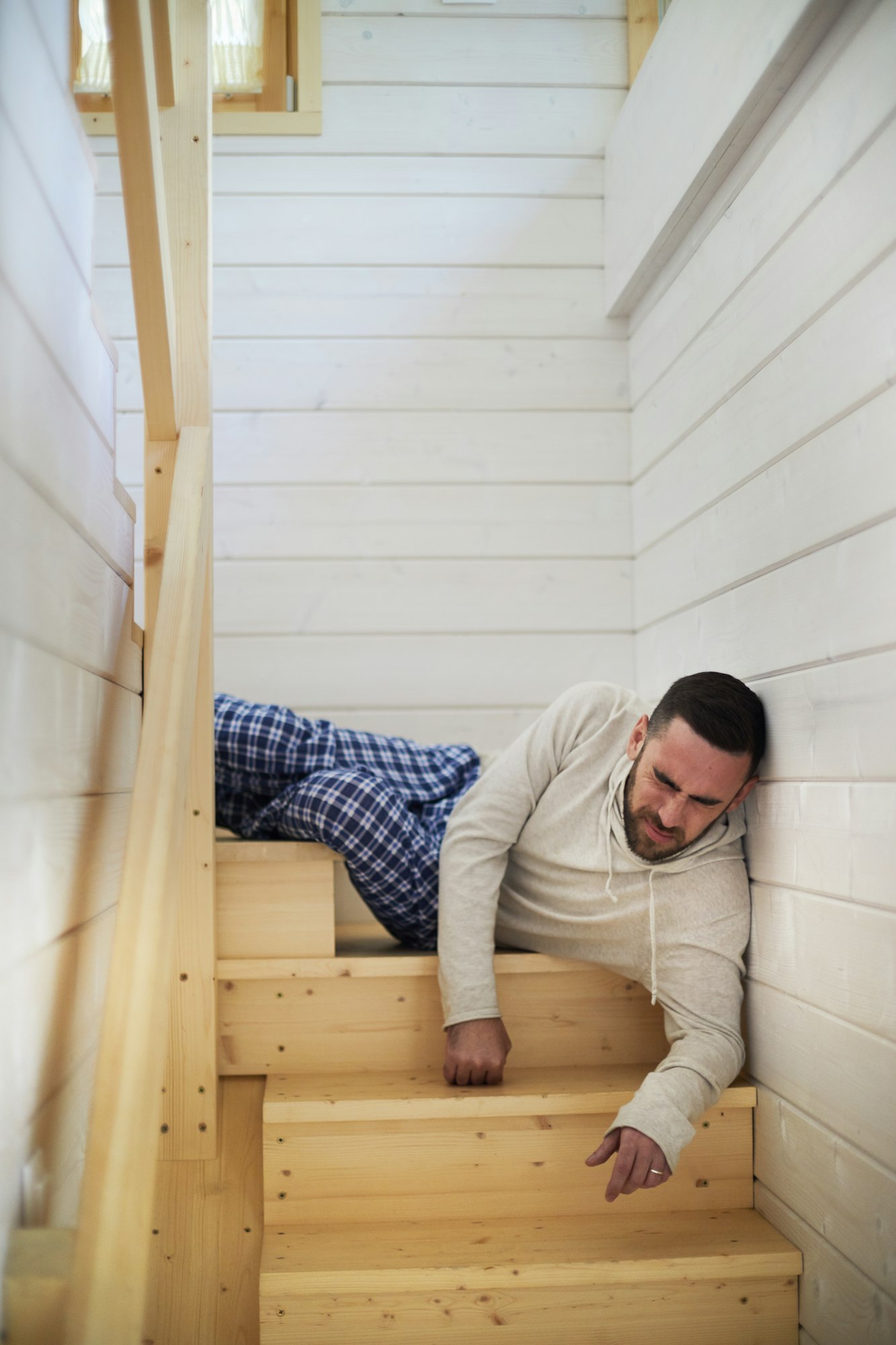 A man in pajamas is lying on wooden stairs and appears to be wincing in pain.