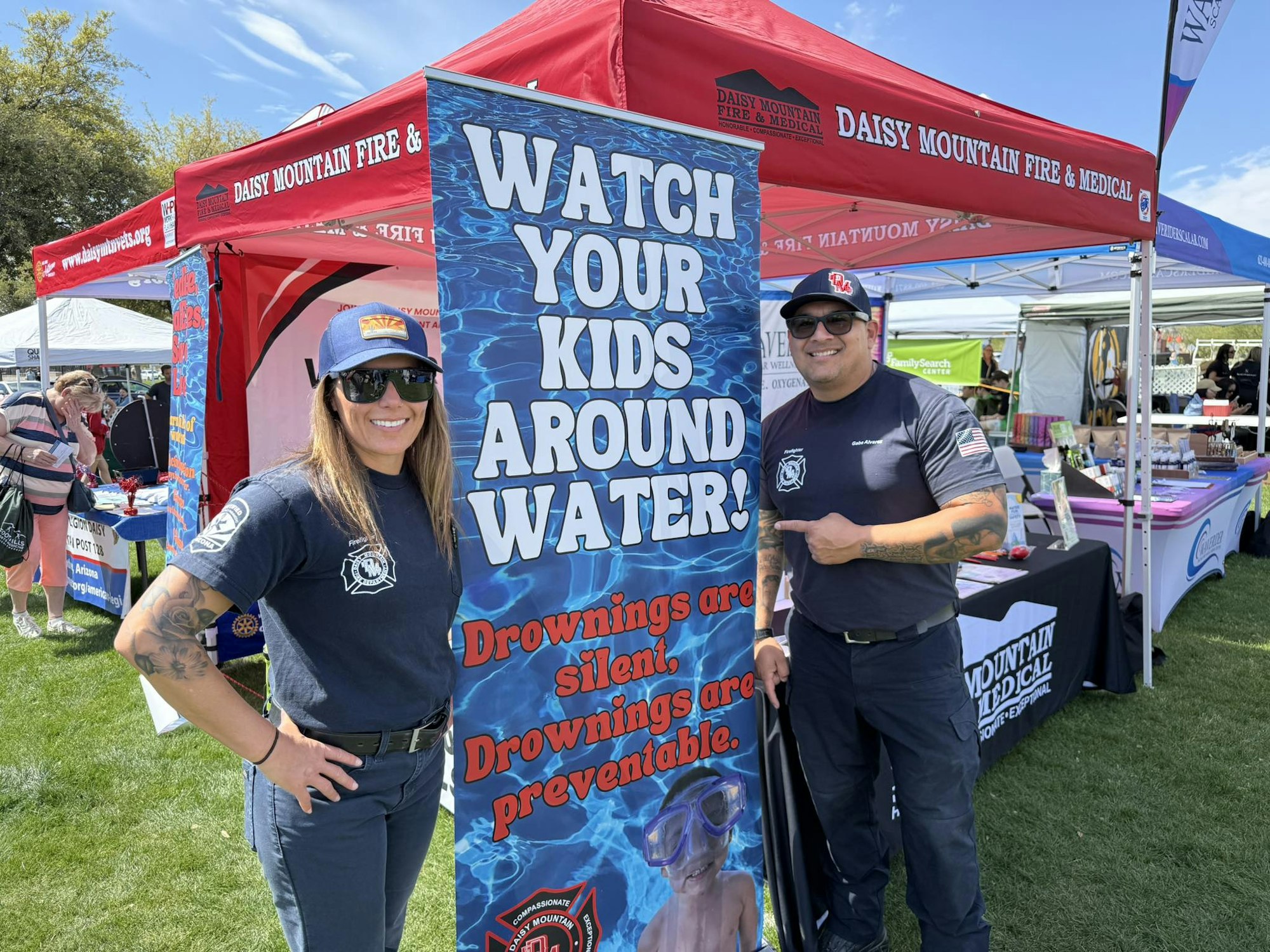 Two people in front of a "Watch Your Kids Around Water" safety poster at a Daisy Mountain Fire & Medical event.